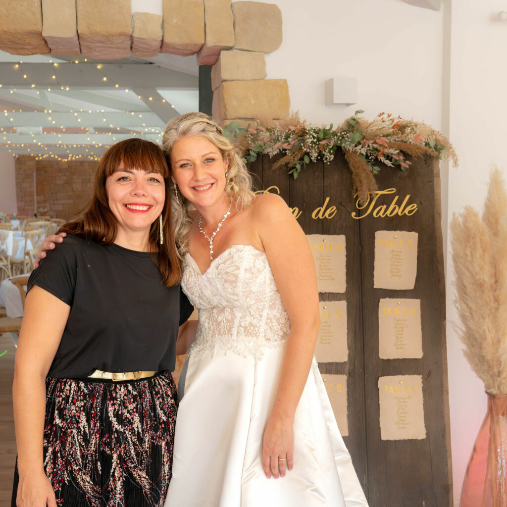 Céline pose à côté de sa cliente en robe de mariée devant le plan de table d'un mariage réalisé par Céline, photo haute luminosité