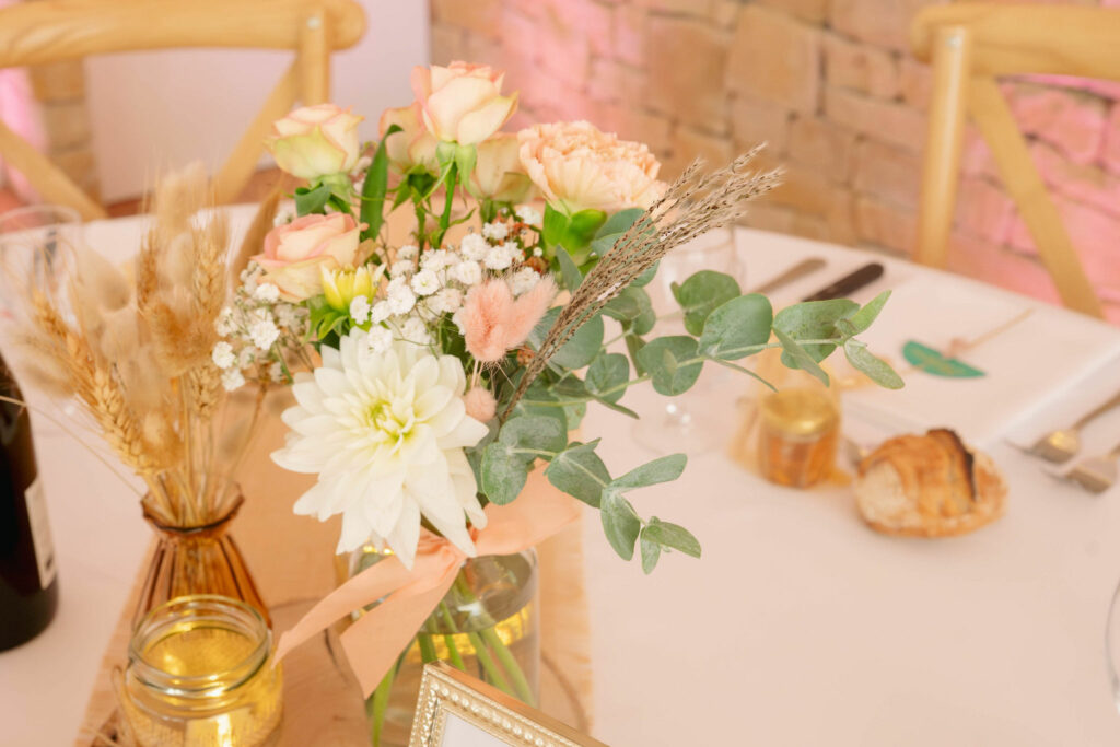 Une table ronde décorée dans des tons beige naturels, bouquets de fleurs séchées au centre de la table, numéro de table gravé sur un rondin de bois par Céline, avec le menu du repas dans un cadre doré, bouteilles de vins