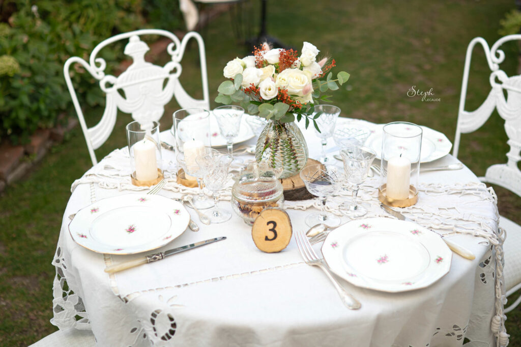 une table est décorée en blanc, avec un bouquet de roses blanches dans un vase au centre, de la vaisselle à fleurs, un rondin de bois avec le numéro gravé par Céline, chaises et nappe élégante,