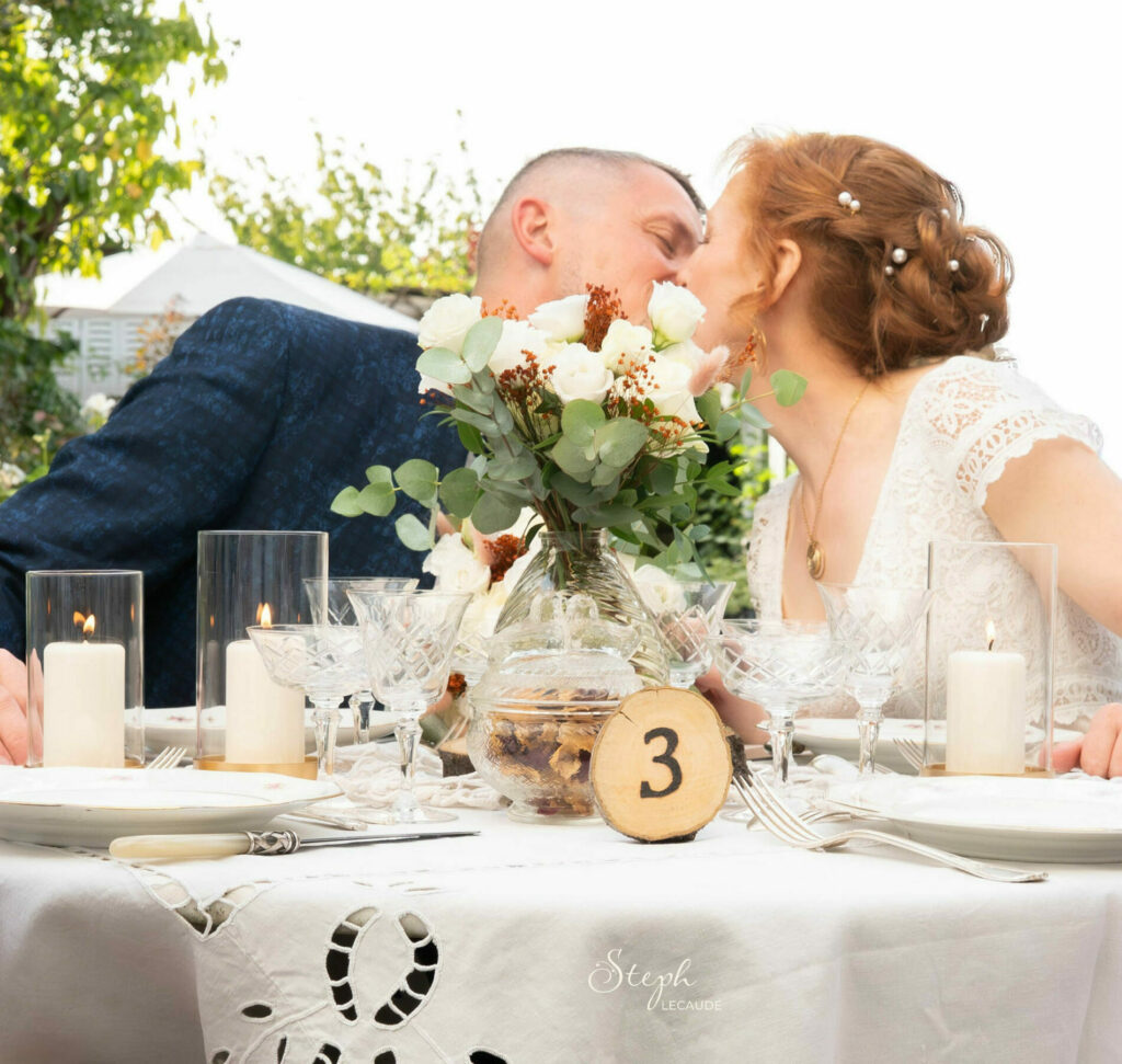 le couple de marié s'embrasse à table, derrière un bouquet de roses blanchesn rondin de bois avec le numéro de la table gravé par Céline, thème retro chic