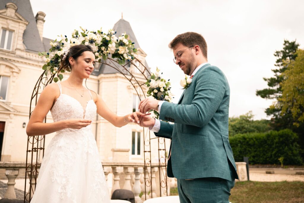 Deux mariés qui se passent la bague au doigts, cérémonie devant un château avec arche de fleurs